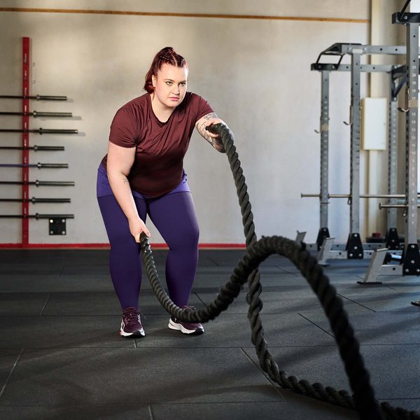 Eine Frau trainiert in einem Fitnessstudio mit Battle Ropes. Sie trägt ein bordeauxfarbenes T-Shirt, lila Leggings und Sportschuhe. Im Hintergrund sind Hanteln an der Wand sowie Krafttrainingsgeräte zu sehen.